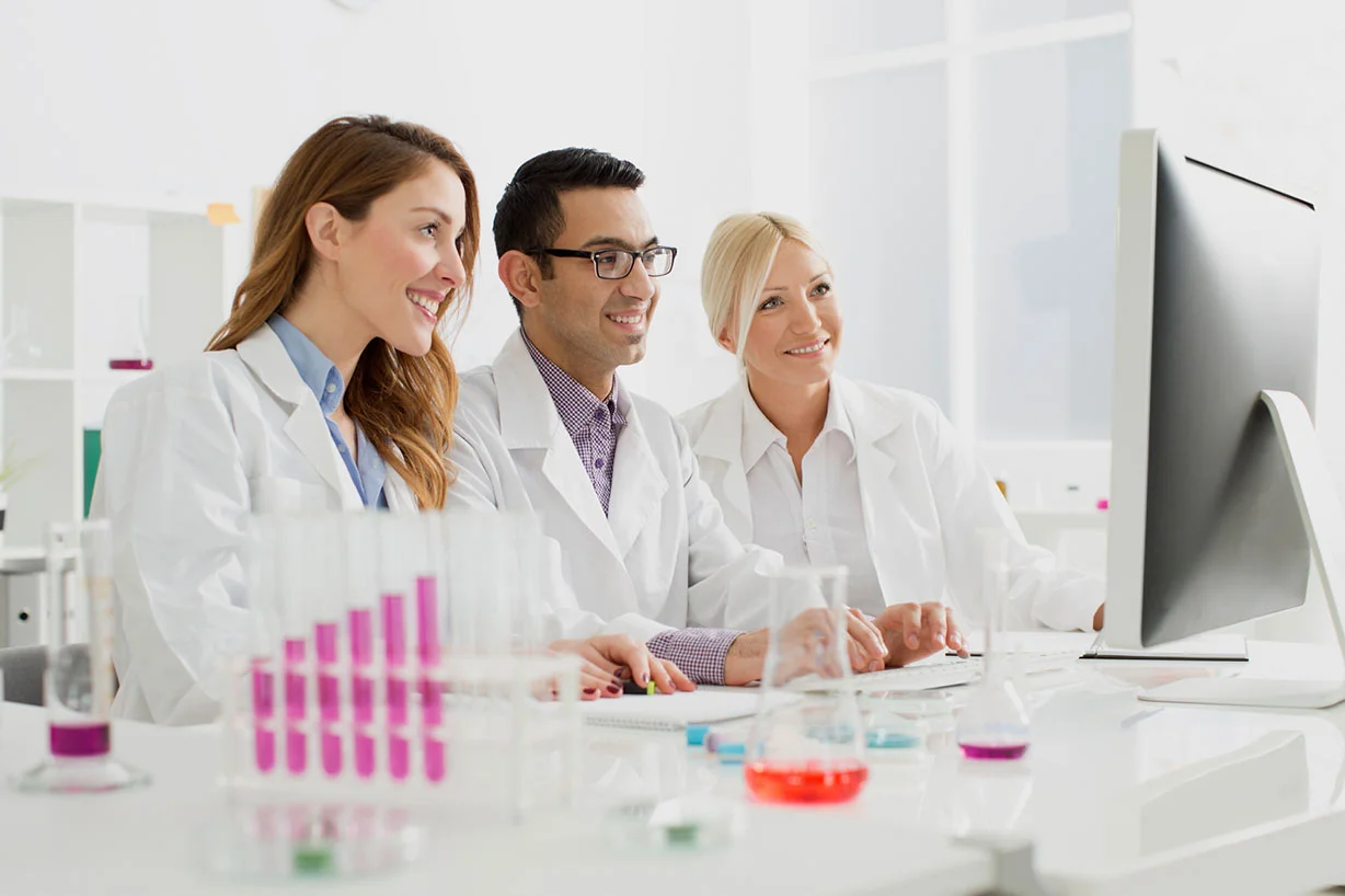 Three smiling scientists in white lab coats, gathered behind a computer screen